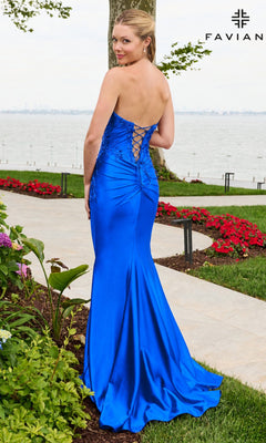 A woman wearing an electric blue Faviana 11647 strapless satin prom dress with floral beaded appliques and a corset-style lace-up back, standing on a paved path outdoors.
