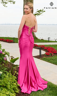 A woman wears a sleeveless, hot pink mermaid-style Faviana 11647 prom dress with floral beaded appliques and a lace-up back, standing on a garden path near the water.