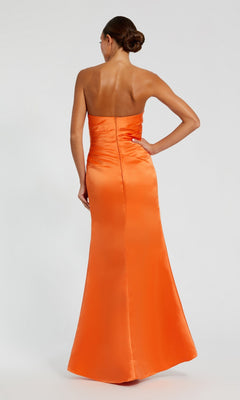 Model in the Mac Duggal 11653 strapless satin prom dress, floor-length and orange, stands with one hand on hip and back to the camera against a plain background.
