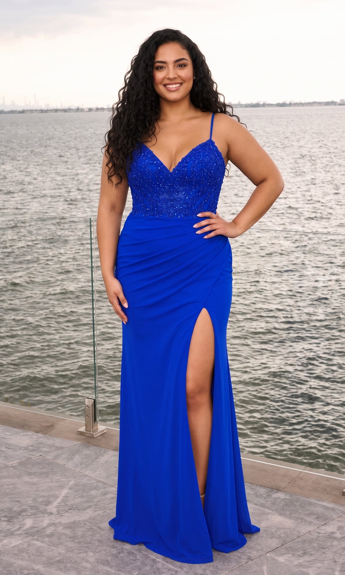 A woman wearing a royal blue Long Plus-Size Prom Dress: Faviana 11653E, a bodycon gown with a beaded bodice and high leg slit, standing by a body of water.
