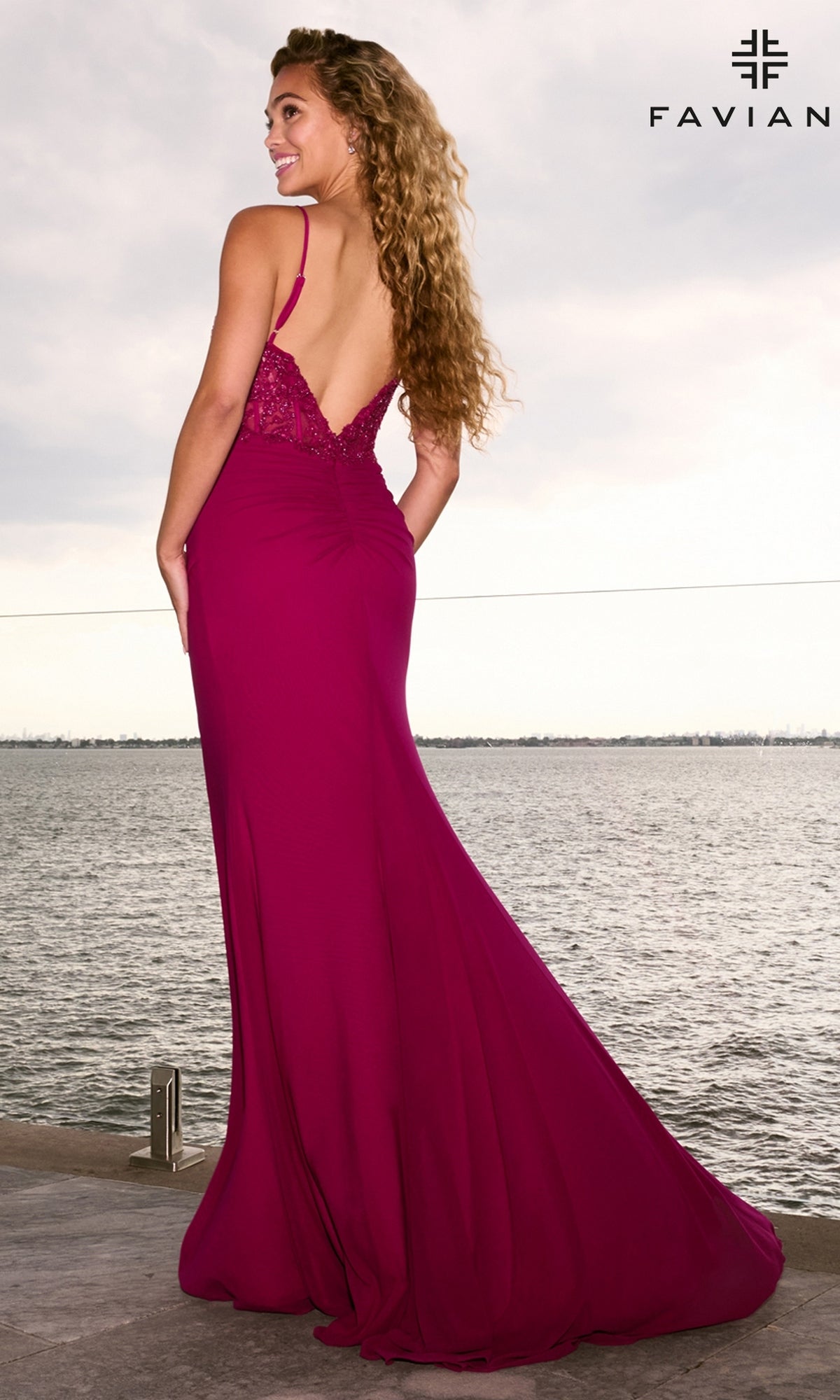 A woman stands by the water in a magenta, floor-length lace corset Faviana 11653 prom dress featuring a beaded, open-back bodice.