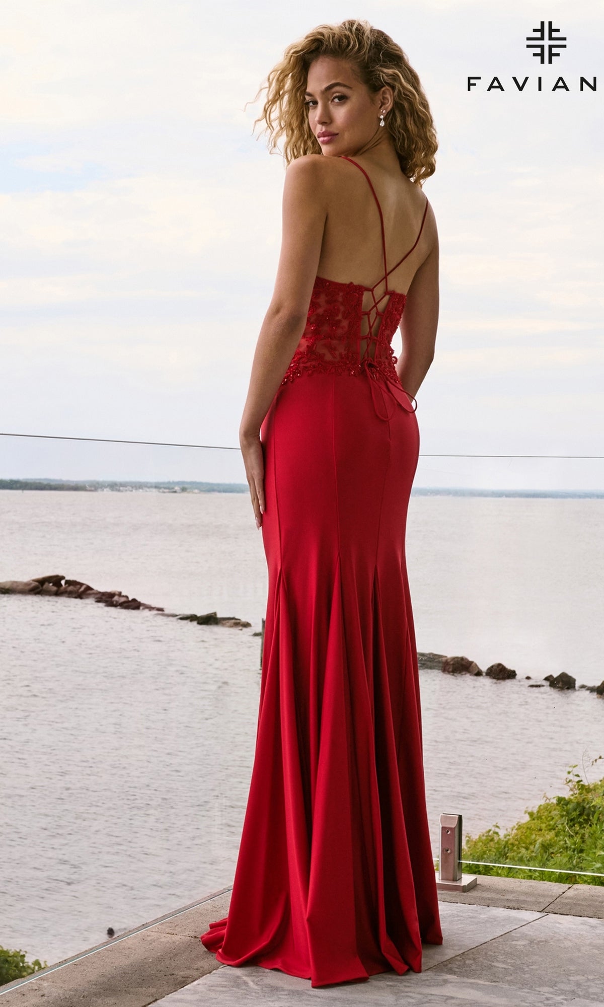 A woman wearing a red floor-length, V-neck mermaid Faviana 11657 prom dress with a lace-up corset back and spaghetti straps, standing on a deck overlooking a body of water.
