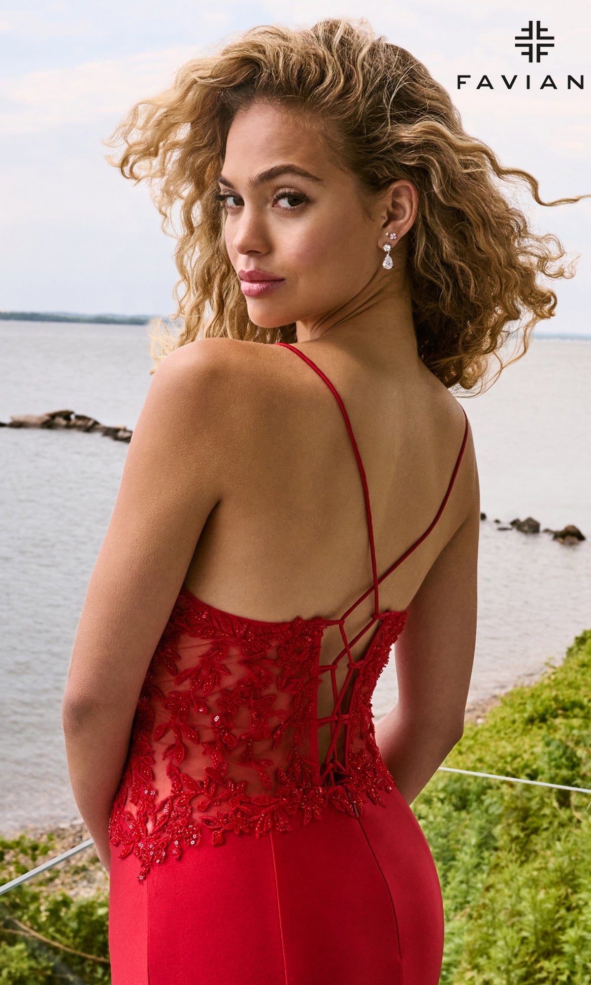 A woman stands near water wearing a Faviana 11657 long prom dress, a floor-length red mermaid gown with a sheer, floral-embroidered V-neck bodice and a lace-up back.