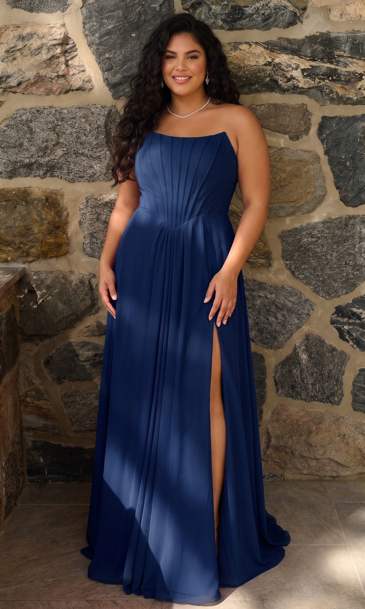 A person wears a navy blue, strapless chiffon Long Plus-Size Prom Dress: Faviana 11672E featuring a floor-length pleated bodice and a high side slit.