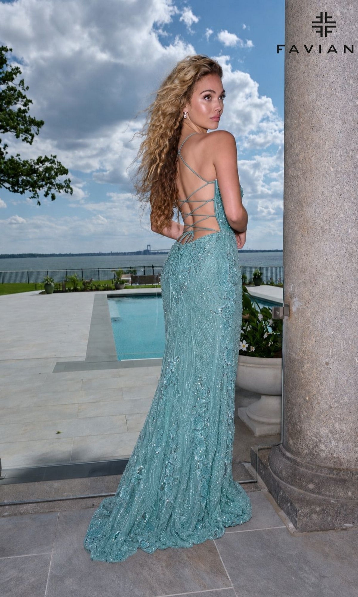 A woman wears a turquoise Faviana 11683 corset prom dress with a lace-up back while standing by a stone pillar overlooking a swimming pool and the ocean.