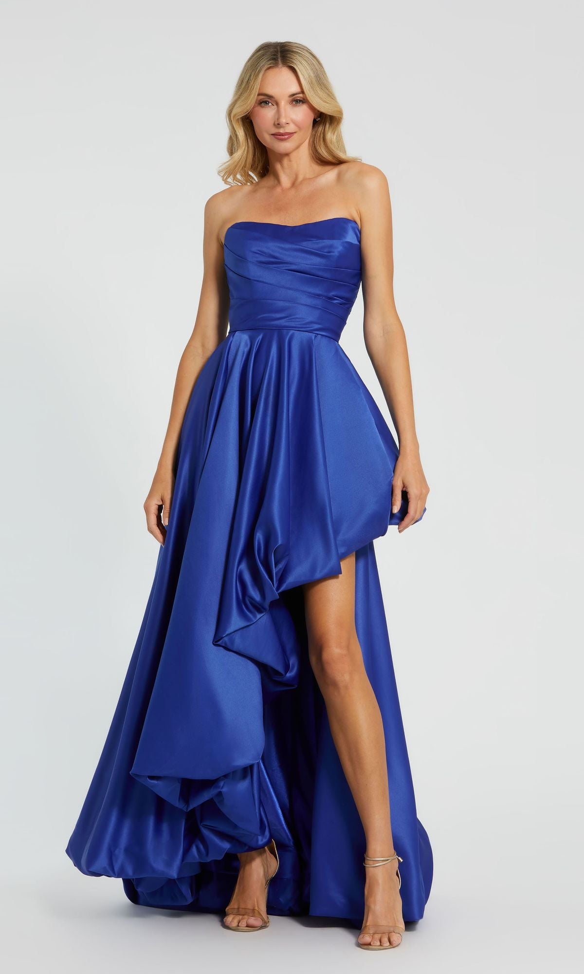 A woman in the Strapless Balloon Prom Dress: Mac Duggal 11685, featuring a royal blue ruched bodice and asymmetrical high-low hem, stands against a plain background.