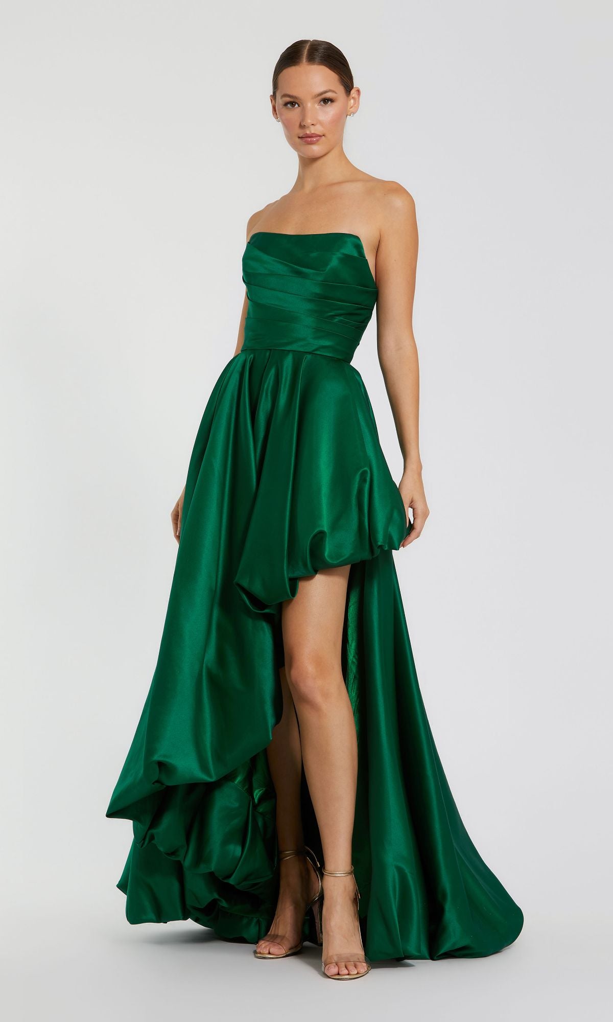 A woman wears the Strapless Balloon Prom Dress: Mac Duggal 11685, an emerald green strapless high-low gown with a ruched bodice and flowing skirt, paired with strappy heels.