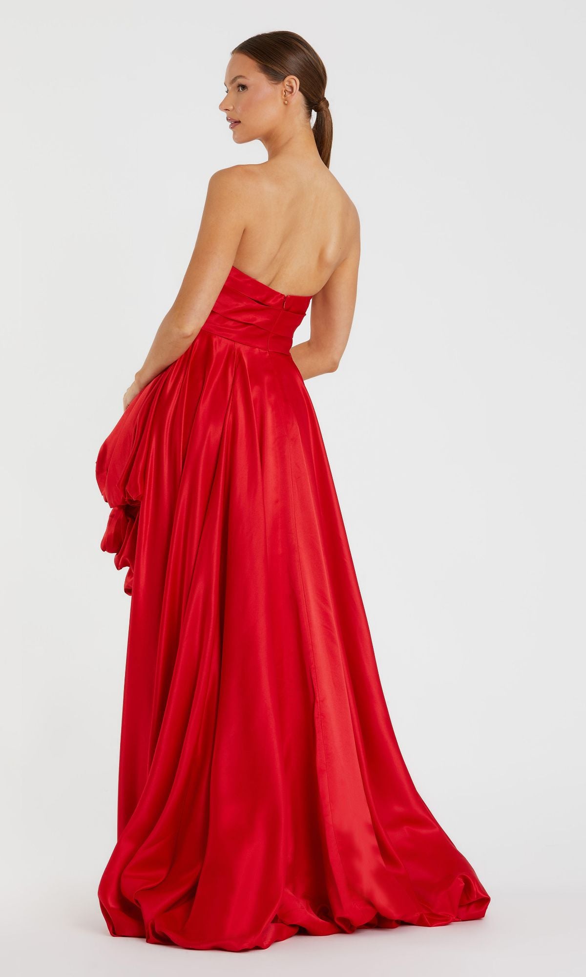A woman in a strapless, floor-length Strapless Balloon Prom Dress (Mac Duggal 11685) in bright red stands facing away, glancing to the side against a plain white background.