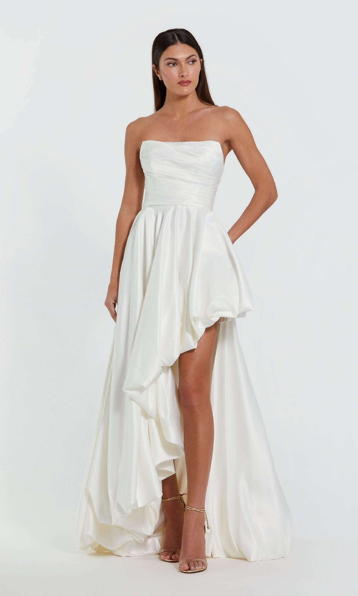 A woman wears the Strapless Balloon Prom Dress by Mac Duggal 11685 in white satin with an asymmetrical high-low hemline and nude heels, standing against a plain white background.