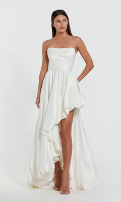 A woman wears the Strapless Balloon Prom Dress by Mac Duggal 11685 in white satin with an asymmetrical high-low hemline and nude heels, standing against a plain white background.