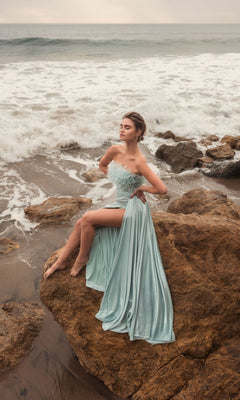 A woman wearing the Long Formal Dress 11690 by Mac Duggal in seafoam green sits on rocks by the shore, with waves and an overcast sky in the background.