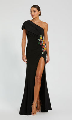 Woman wearing the Mac Duggal 11769 Long Formal Dress in black, showcasing a one-shoulder gown with a thigh-high slit and vibrant 3D floral applique embroidery at the waist, standing against a plain background.