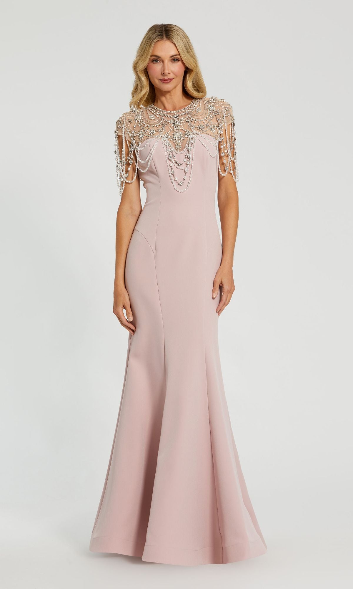 A woman stands against a white background in the Mac Duggal 11775 Pearl-Neckline Long Formal Dress, a floor-length pale pink gown featuring an embellished beaded cape overlay on the shoulders and chest.