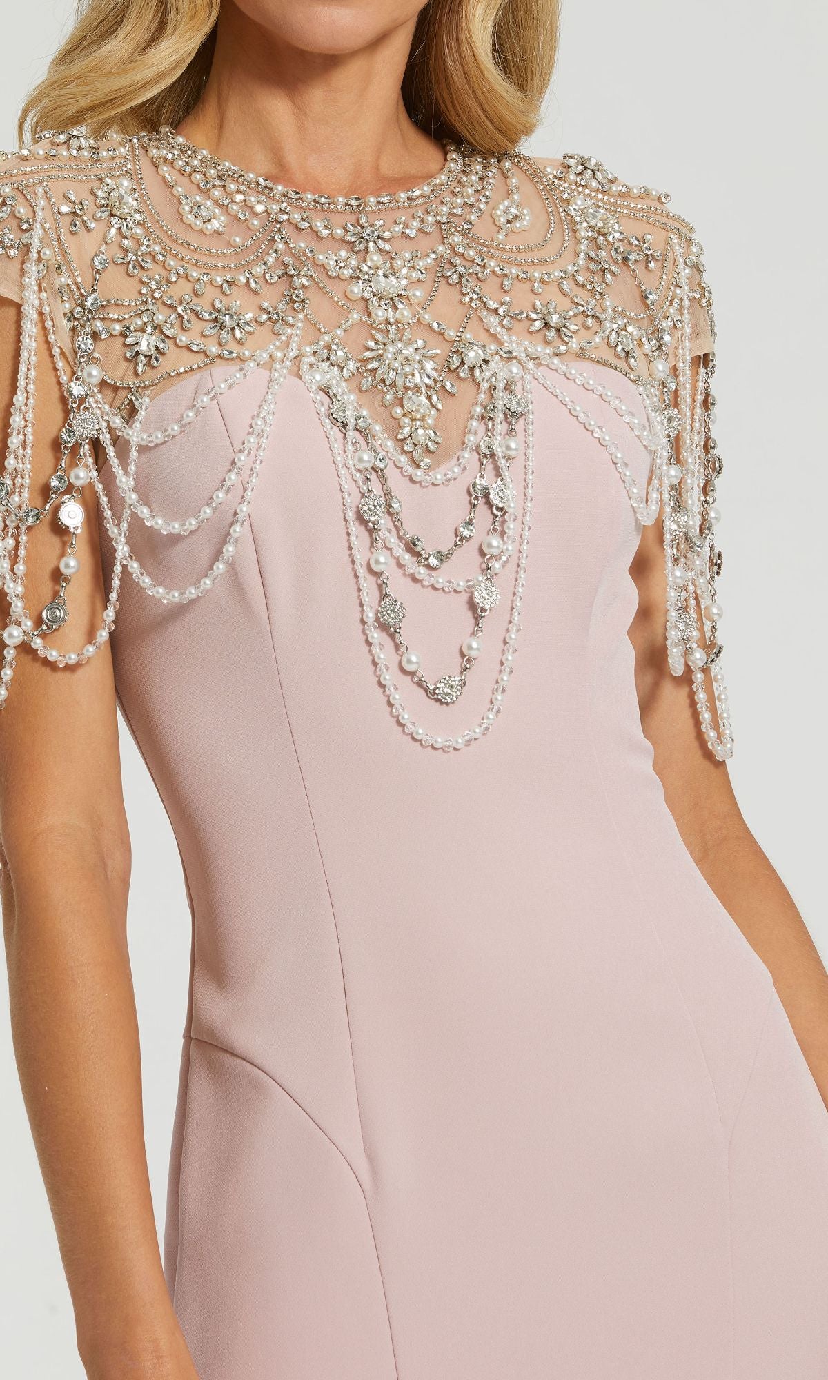A woman wearing the Mac Duggal 11775 Pearl-Neckline Long Formal Dress, featuring a pale pink hue and an ornate beaded overlay draped elegantly over her shoulders and upper chest.