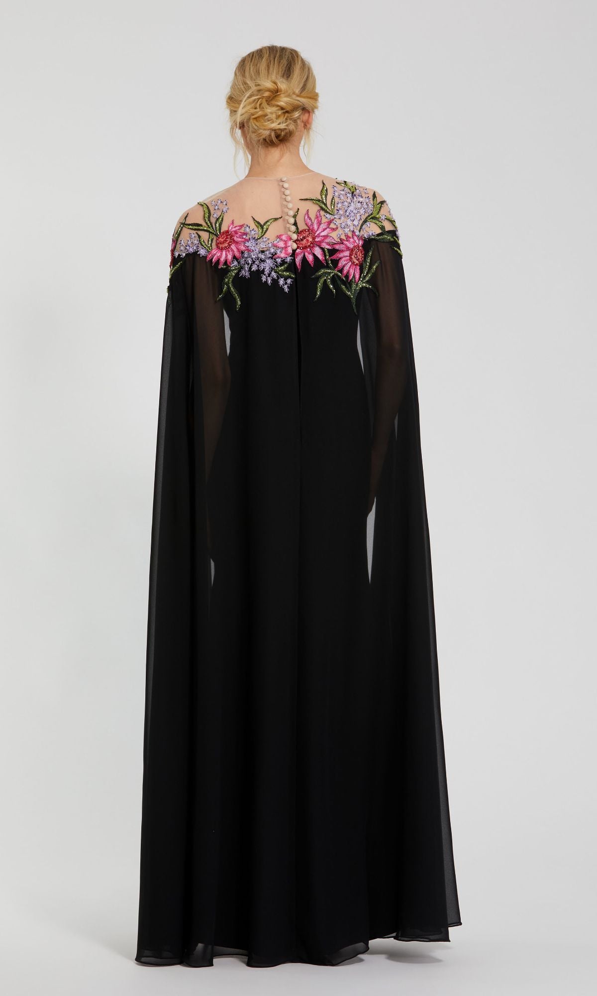 A woman with blonde hair in an updo models the Mac Duggal 11798 long formal evening gown, featuring black fabric, sheer cape sleeves, and beaded floral embroidery across the shoulders, photographed from behind against a plain background.