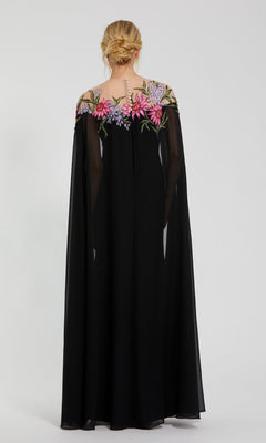 A woman with blonde hair in an updo models the Mac Duggal 11798 long formal evening gown, featuring black fabric, sheer cape sleeves, and beaded floral embroidery across the shoulders, photographed from behind against a plain background.