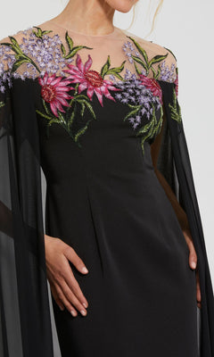 A person wears the Mac Duggal 11798 long formal evening gown, featuring sheer sleeves and vibrant beaded floral embroidery on the upper bodice and shoulders.