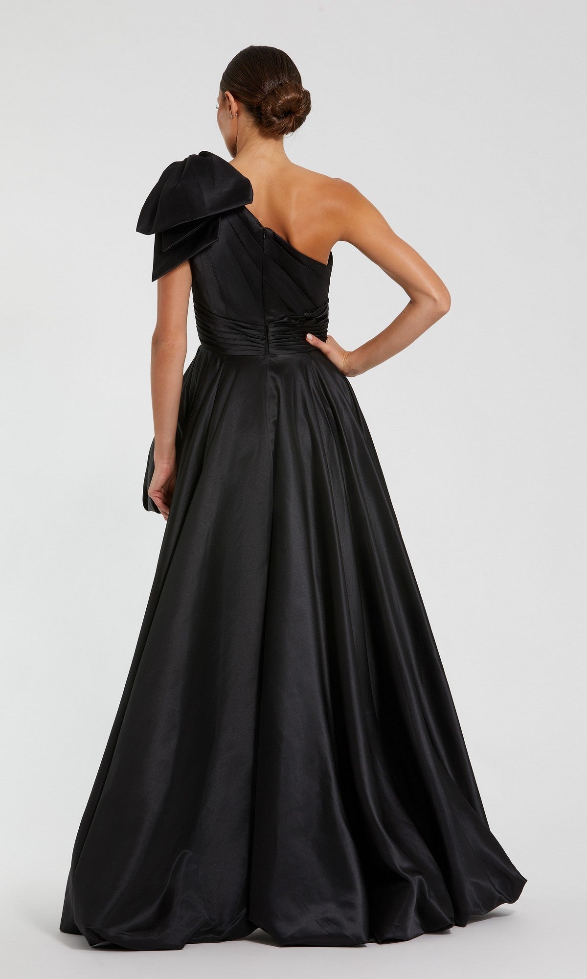 A woman stands with her back to the camera, wearing the Mac Duggal 11800 Bow Long Bubble Prom Dress—an elegant black one-shoulder gown with a large bow accent, set against a plain background.