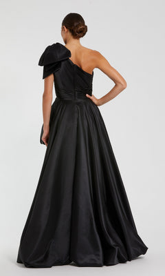 A woman stands with her back to the camera, wearing the Mac Duggal 11800 Bow Long Bubble Prom Dress—an elegant black one-shoulder gown with a large bow accent, set against a plain background.
