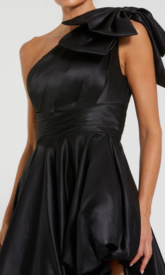 A woman wears the Mac Duggal 11800 Bow Long Bubble Prom Dress, featuring black satin, an asymmetrical one-shoulder design with a large shoulder bow, and a sleek, modern silhouette.