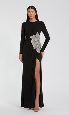 A woman stands facing forward in the Mac Duggal 11809 long sleeve black formal dress, featuring a high slit and a large silver floral embellishment at the waist.
