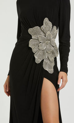 A person wearing the Mac Duggal 11809 long sleeve black formal dress featuring a high slit and a large silver beaded floral embellishment at the waist.