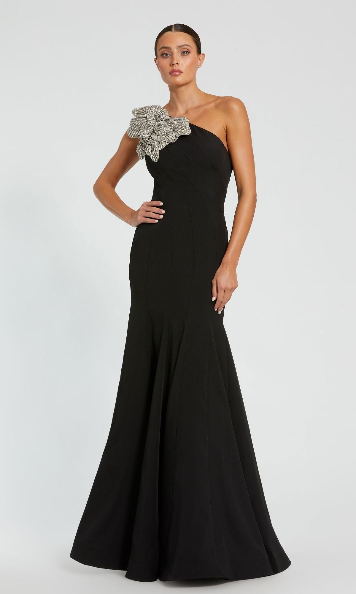 A woman in the Mac Duggal 11810 Beaded-One-Shoulder Prom Gown, featuring a black design with a large silver floral embellishment on the shoulder, stands against a plain light background.