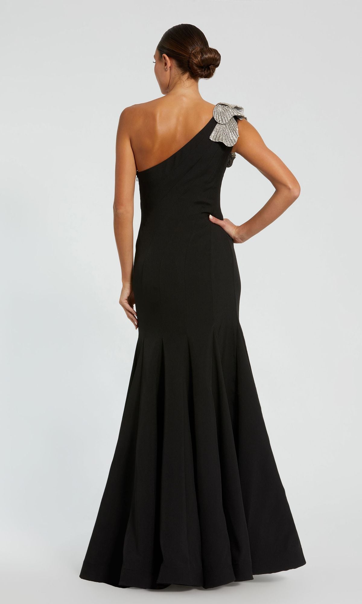 A woman stands facing away, hand on hip, in the Mac Duggal 11810 Beaded-One-Shoulder Prom Gown, a black floor-length dress with a silver bow accent on the shoulder.