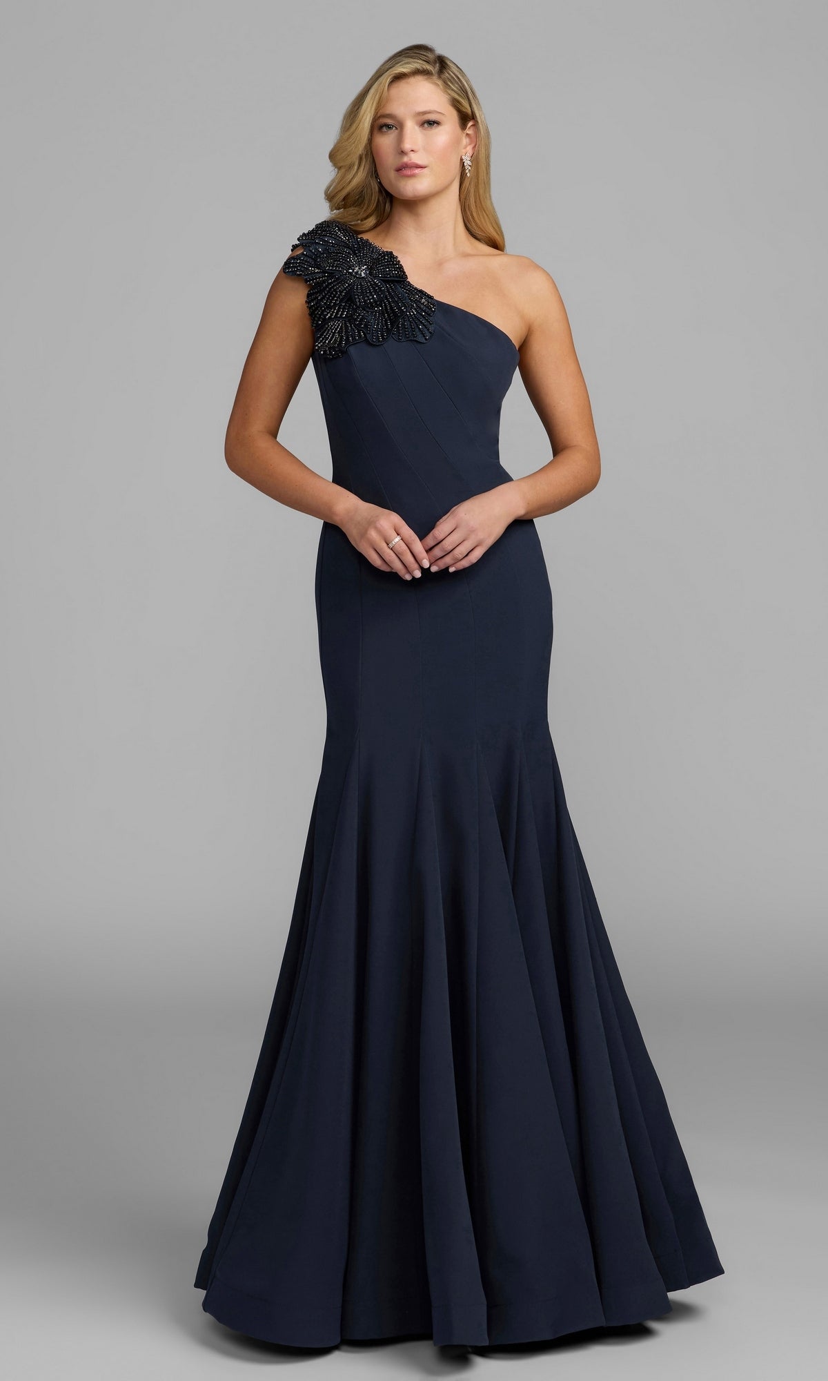 A woman stands against a plain gray background wearing the Beaded-One-Shoulder Prom Gown: Mac Duggal 11810 in Midnight—a floor-length, one-shoulder evening gown featuring black beaded embellishment on the shoulder. This striking dress exudes timeless sophistication.
