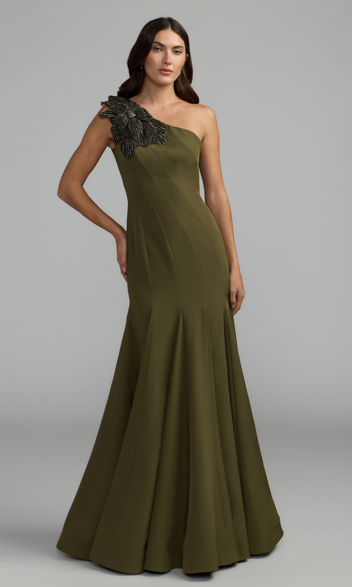 A woman is wearing the Beaded-One-Shoulder Prom Gown: Mac Duggal 11810 in Olive, which features black beaded embellishment on the single shoulder, as she stands against a plain grey background.