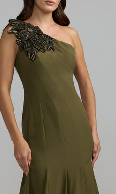 A woman wearing the Beaded-One-Shoulder Prom Gown: Mac Duggal 11810 in olive green, featuring a dramatic black beaded floral embellishment on the shoulder, evokes the elegance of a signature Mac Duggal formal evening gown.