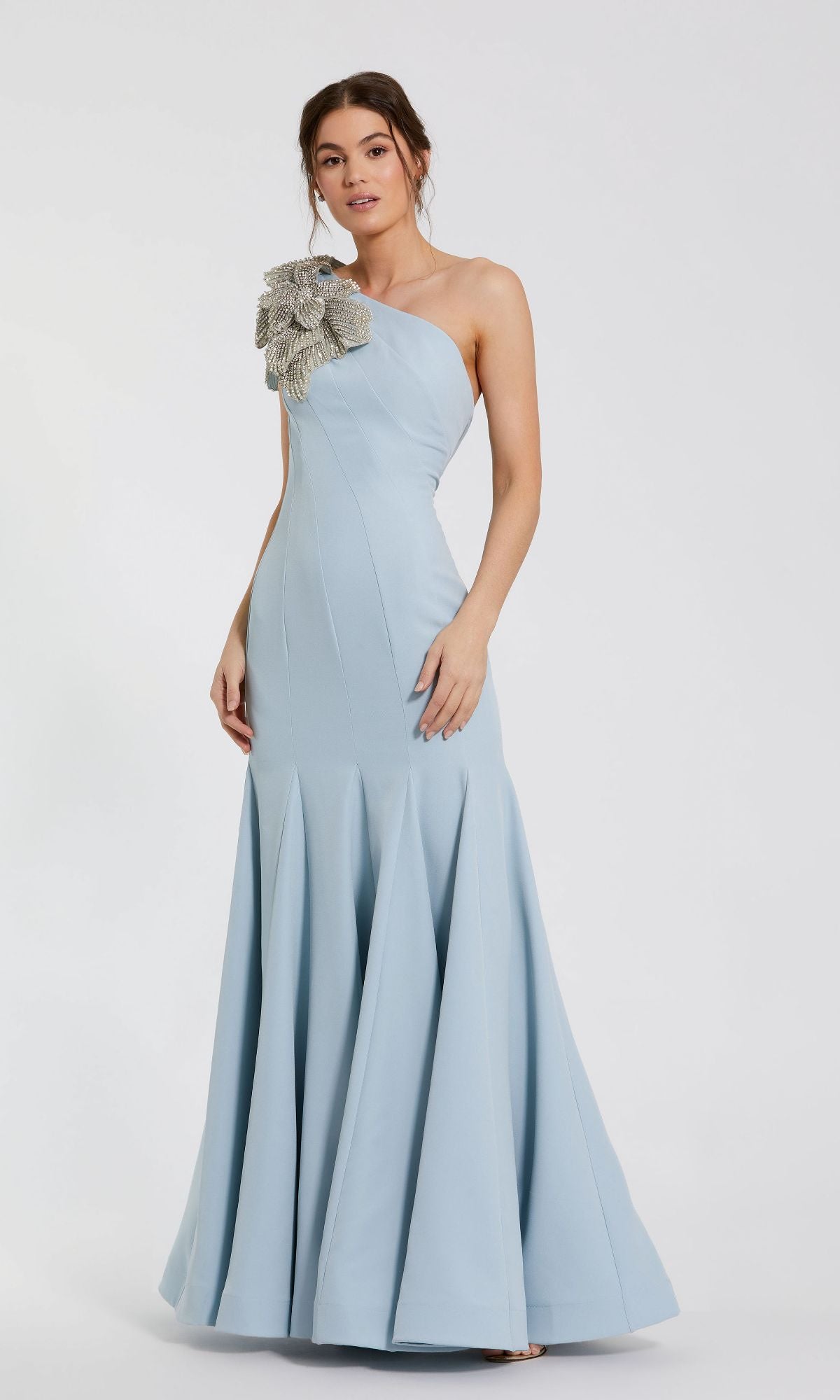 A woman poses against a plain white background in the Mac Duggal 11810 Beaded-One-Shoulder Prom Gown, a light blue dress featuring a prominent silver floral embellishment on one shoulder.