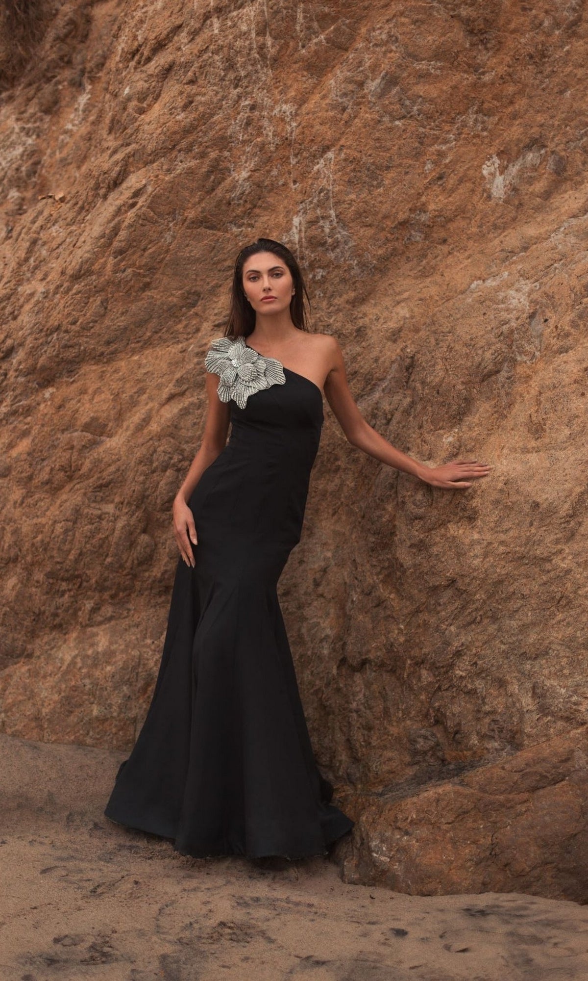 A woman in the Mac Duggal 11810 Beaded One-Shoulder Prom Gown with a large silver accessory stands against a rocky outdoor wall.