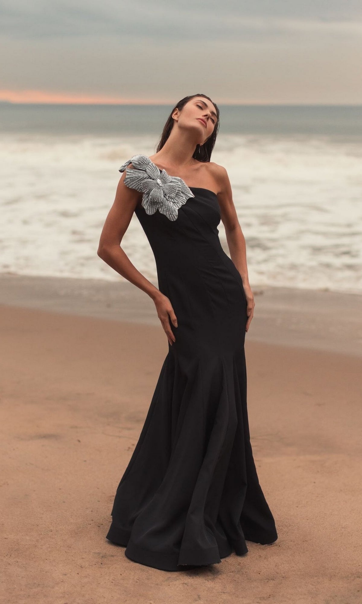 A woman stands on a beach in the Mac Duggal 11810 Beaded-One-Shoulder Prom Gown—a long black dress featuring a dramatic silver floral embellishment on one shoulder.