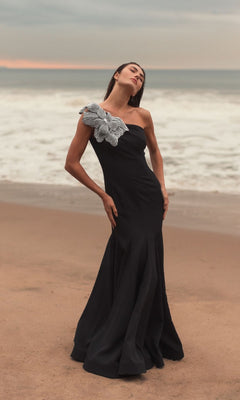 A woman stands on a beach in the Mac Duggal 11810 Beaded-One-Shoulder Prom Gown—a long black dress featuring a dramatic silver floral embellishment on one shoulder.