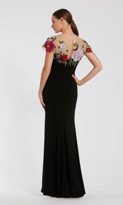 A woman stands with her back to the camera, one hand on her hip, wearing the Mac Duggal 11817 Floral Long Black Formal Dress featuring a sheer, floral-embroidered top.
