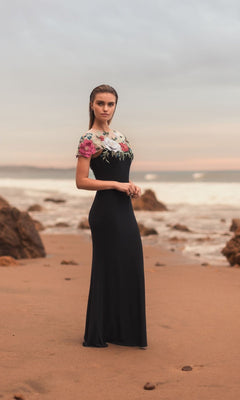Wearing the Mac Duggal 11817 Floral Long Black Formal Dress, a woman stands on a sandy beach as her embroidered gown flows in the sunset by the ocean and rocky shore.