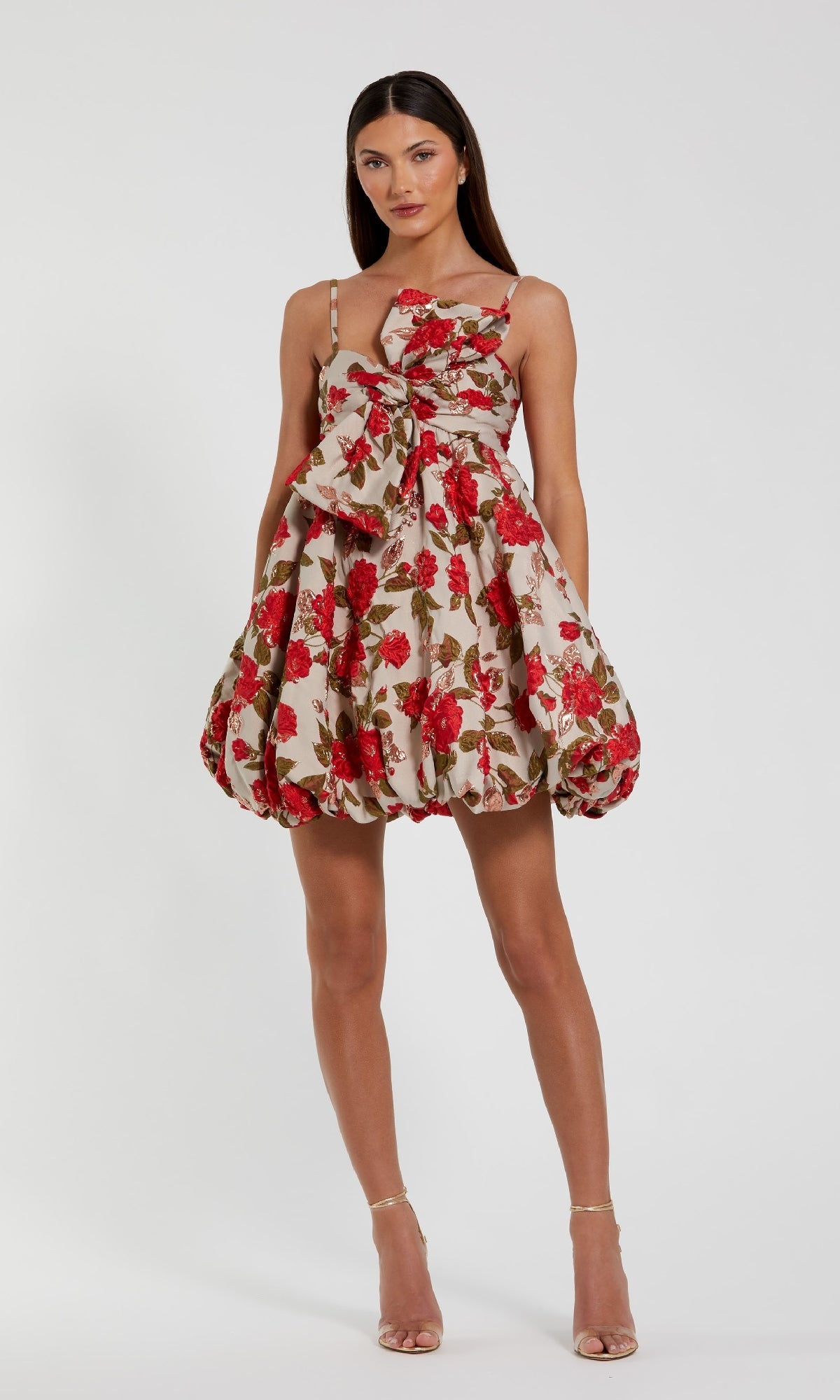 A woman models the Mac Duggal 11822 Short Bubble-Hem Hoco Dress, a sleeveless floral-print design with a large front bow, standing against a plain white background.