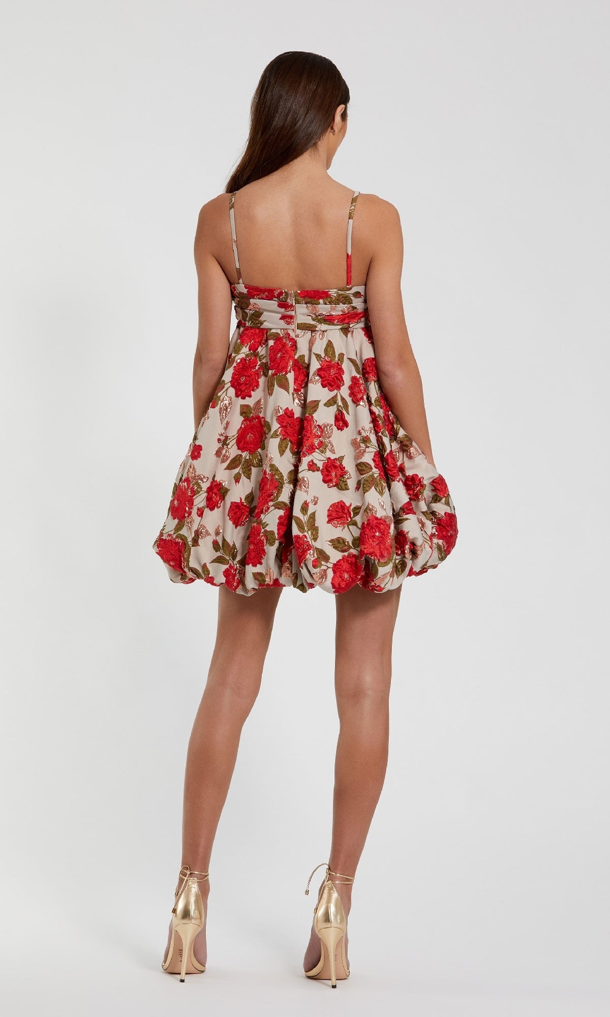 A woman faces away in the Mac Duggal 11822 Short Bubble-Hem Hoco Dress, a sleeveless beige mini with red floral prints, styled with gold high heels against a plain white background.