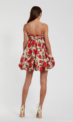 A woman faces away in the Mac Duggal 11822 Short Bubble-Hem Hoco Dress, a sleeveless beige mini with red floral prints, styled with gold high heels against a plain white background.