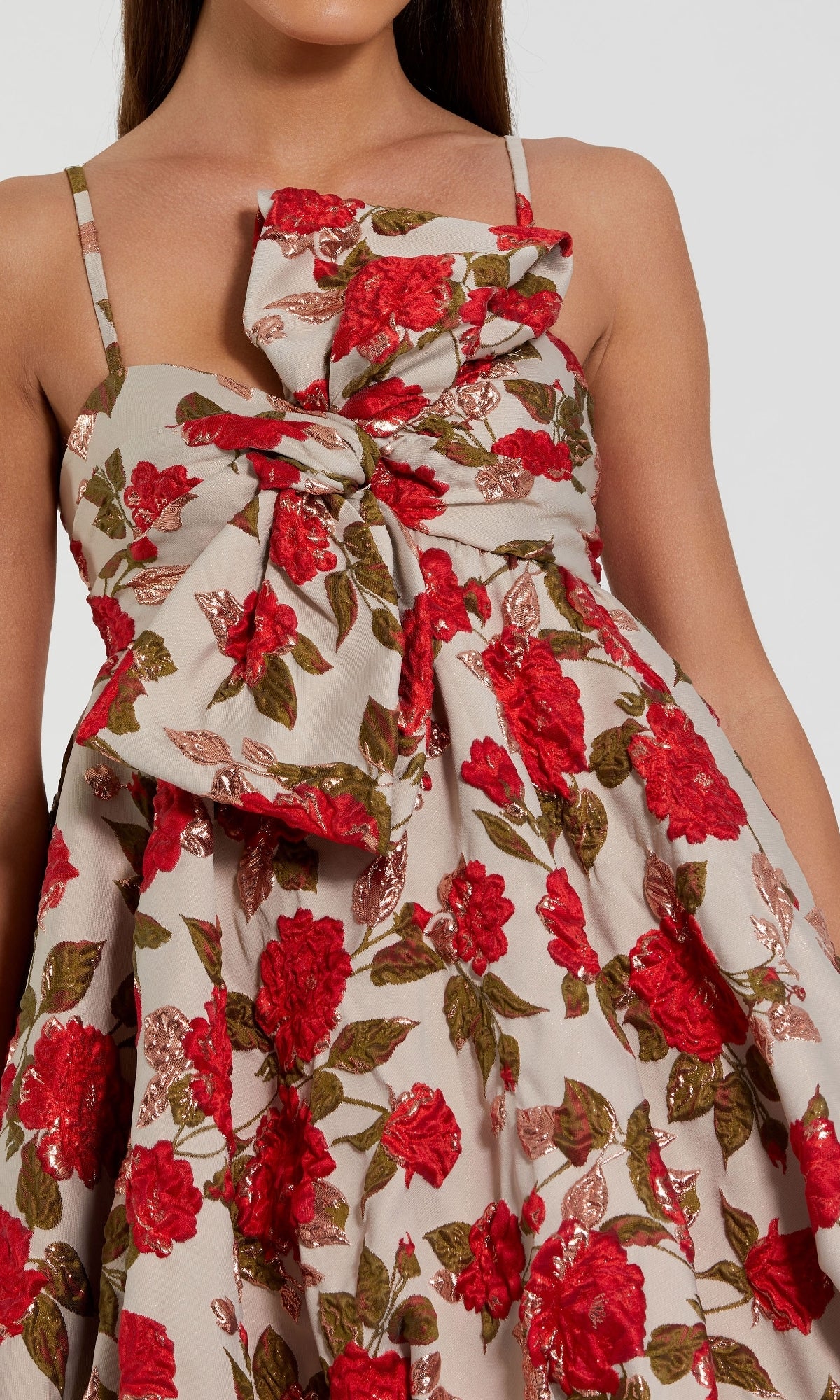 A woman wears the Mac Duggal 11822 short bubble-hem hoco dress with thin straps, a large front bow, and a red and green floral print on a beige background.