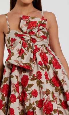 A woman wears the Mac Duggal 11822 short bubble-hem hoco dress with thin straps, a large front bow, and a red and green floral print on a beige background.