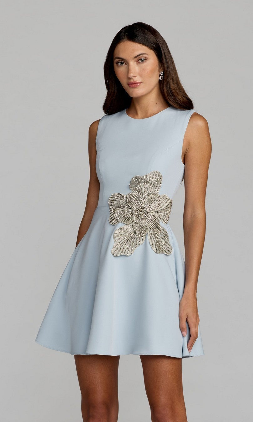 A woman stands against a plain background wearing the Mac Duggal 11829 Short A-Line Cocktail Dress in light blue, detailed with a large metallic floral embellishment at the waist.