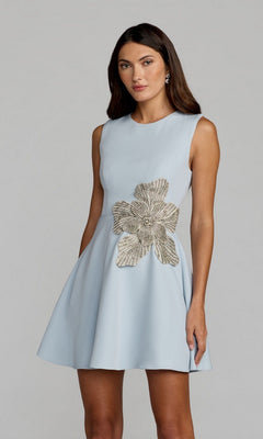A woman stands against a plain background wearing the Mac Duggal 11829 Short A-Line Cocktail Dress in light blue, detailed with a large metallic floral embellishment at the waist.