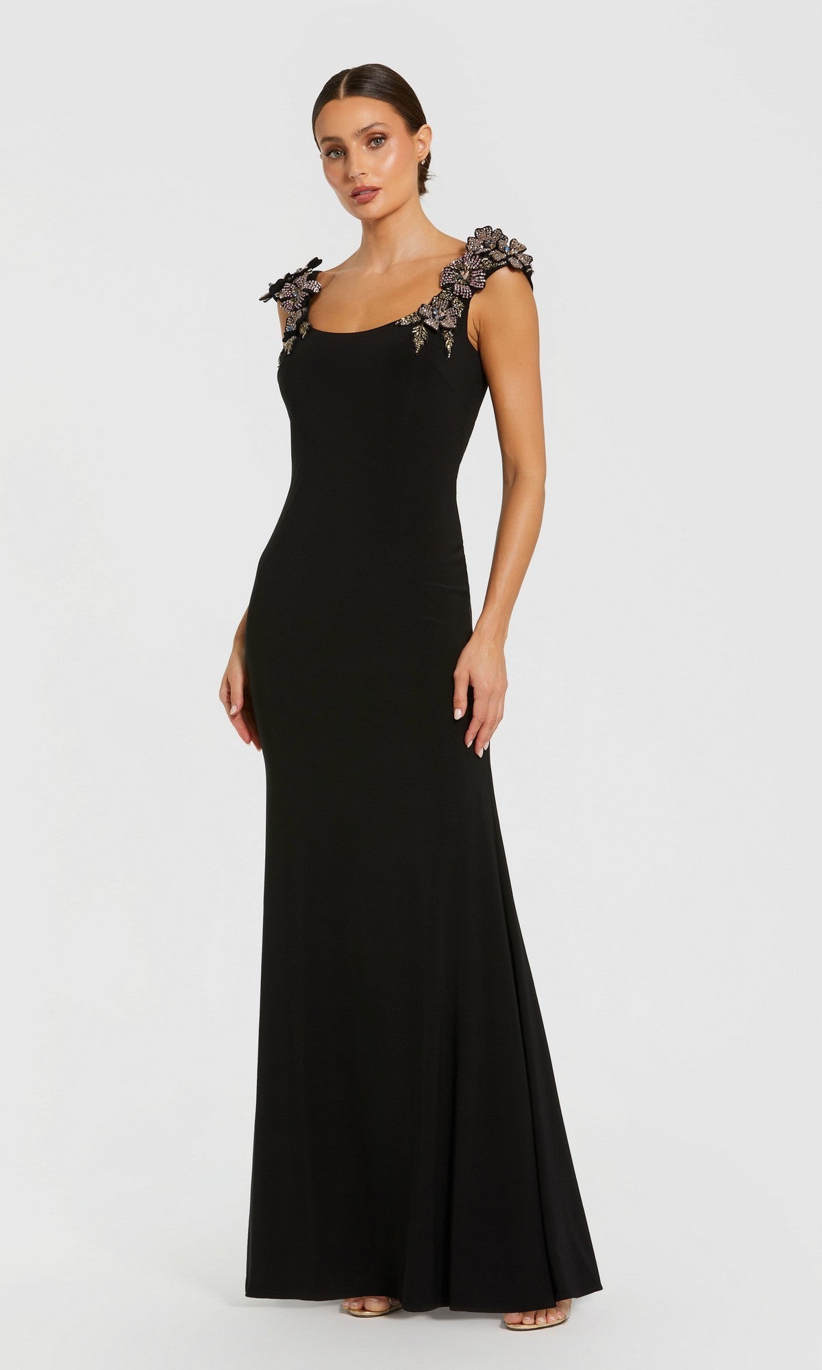 A woman poses against a plain white background wearing the Mac Duggal 11842 long formal dress in black, featuring a fitted silhouette and decorative 3D floral embellishments on the shoulders.
