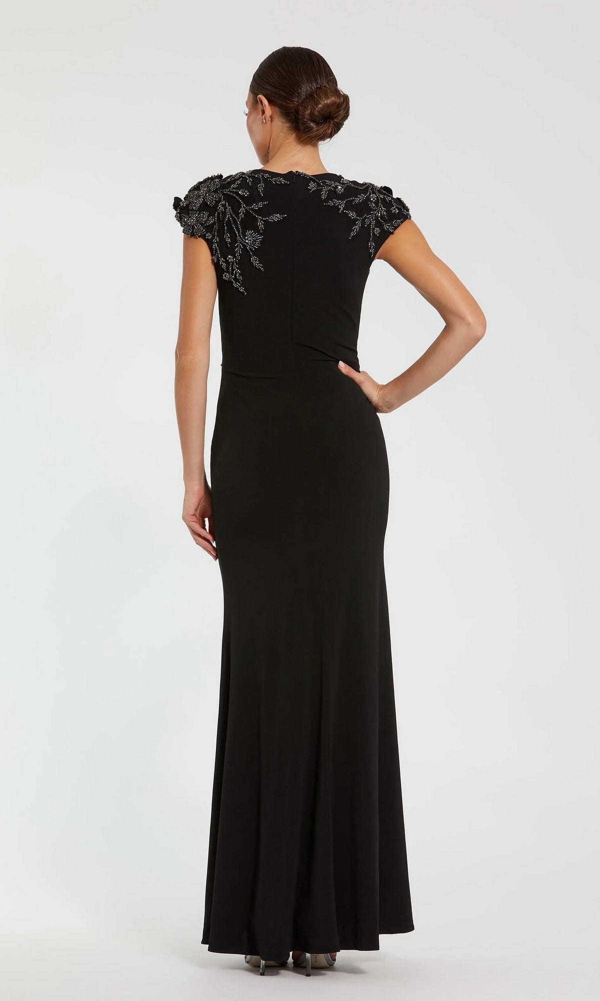 A woman wearing the Mac Duggal 11847 long formal dress, a black column style with short sleeves and silver shoulder embellishments, stands facing away from the camera against a plain background.
