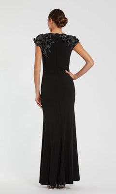 A woman wearing the Mac Duggal 11847 long formal dress, a black column style with short sleeves and silver shoulder embellishments, stands facing away from the camera against a plain background.