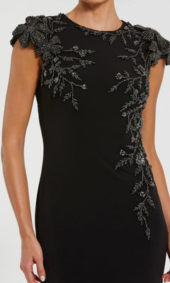 A woman models the Mac Duggal 11847 long formal dress, featuring a black column silhouette with detailed bead and leaf embellishments on the sleeves and upper bodice, set against a plain background.