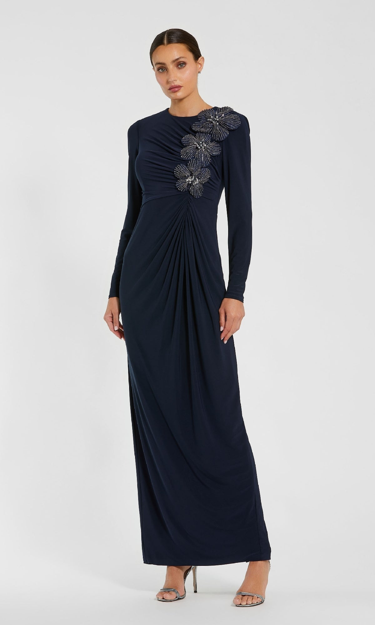 A woman stands against a plain light background wearing the Mac Duggal 11850, a dark blue long formal dress with long sleeves and floral shoulder embellishments, paired with silver high-heeled sandals.