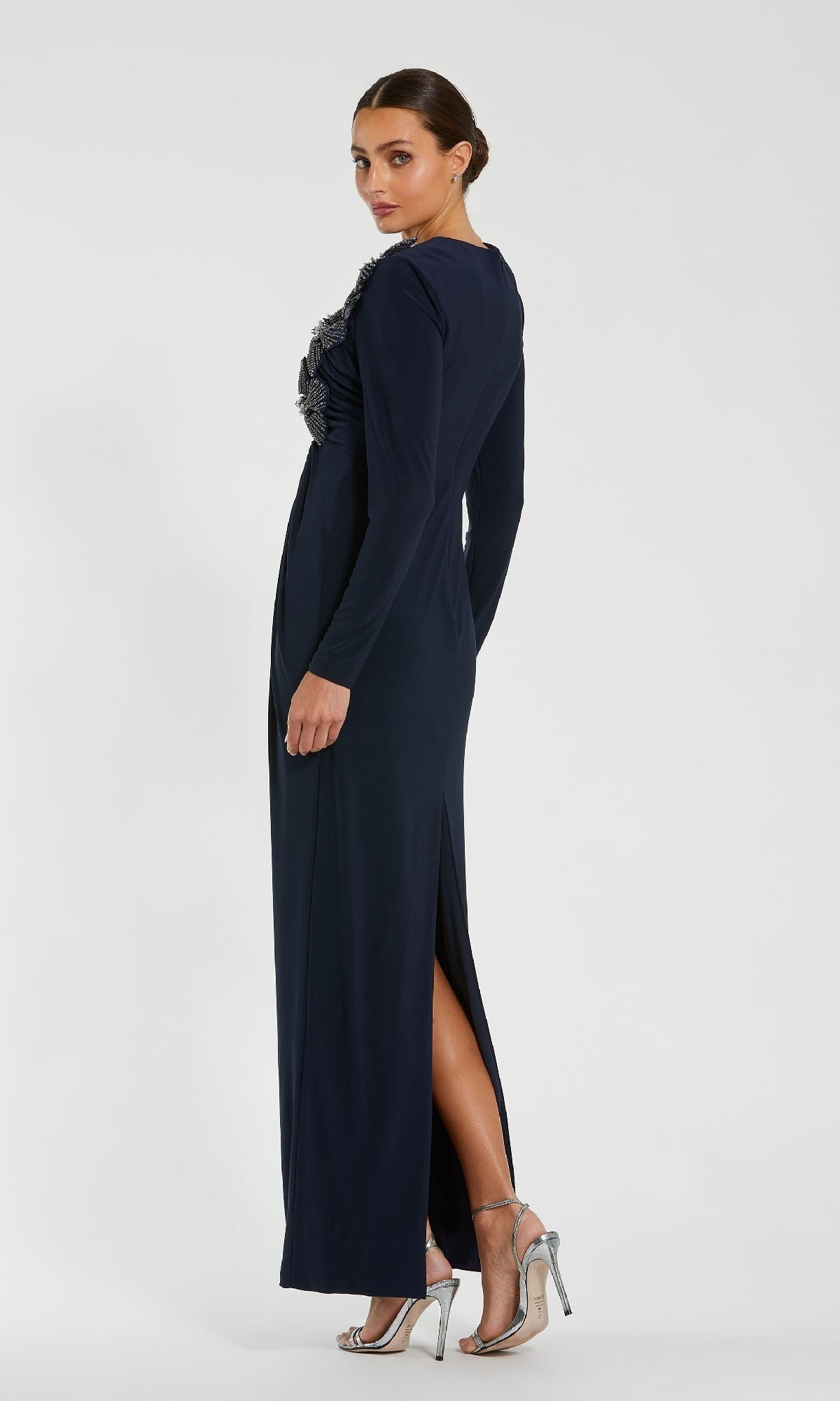Woman in a Mac Duggal 11850 dark blue long formal dress with long sleeves and side slit, wearing silver high-heeled sandals and her hair pulled back, standing against a plain white background.